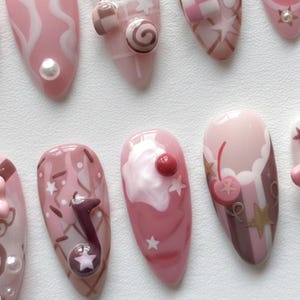 May include: A collection of pink and white nail art designs. The nails feature various designs, including swirls, candies, cherries, and musical notes. Some nails have 3D embellishments, and others have star and stripe patterns.