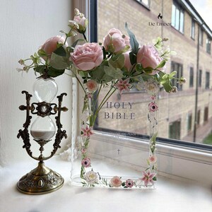 Puede incluir: Un reloj de arena ornamentado con acabado bronceado se encuentra junto a un marco acrílico transparente con la inscripción "HOLY BIBLE". El marco está decorado con detalles florales rosas y blancos, y un ramo de rosas rosas se encuentra encima.