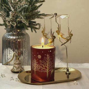 May include: A red candle in a glass jar with a gold tree and deer design. A gold fairy carousel spins above the candle on a gold tray. A vase with greenery is in the background.