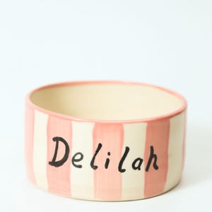 K&ouml;nnte beinhalten: Eine Keramik-Haustier-Sch&uuml;ssel mit cremefarbenem Innenraum und einem rosa-cremefarbenen gestreiften &Auml;u&szlig;eren. Der Name "Delilah" ist in schwarzer Schrift auf die Seite geschrieben. Der Rand ist rosa bemalt.