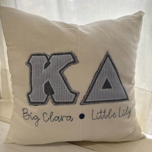 Pode incluir: Almofada quadrada creme com as letras "KA" aplicadas em tecido listrado azul e branco. Abaixo das letras, as palavras "Big Clara" e "Little Lily" são bordadas em itálico.