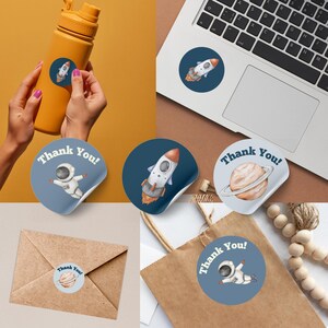 May include: A collection of round stickers with space-themed illustrations. Designs include a rocket ship, an astronaut, and a planet with rings. Some stickers have the text "Thank You!". The stickers are shown on a water bottle, laptop, and gift bag.