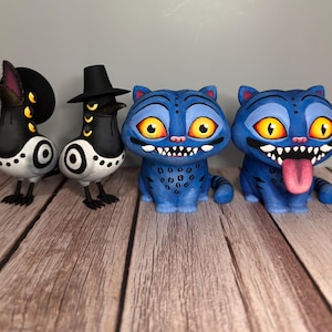 May include: A collection of whimsical, hand-painted figurines. Two bird-like figures in black and white with hats, and two blue cat-like creatures with large yellow eyes and open mouths. The cat figures have visible teeth and one has its tongue out.