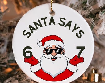 May include: White ceramic Christmas ornament with the text "SANTA SAYS" and a cartoon Santa Claus wearing sunglasses. The numbers 6 and 7 are on either side of Santa. The ornament hangs from a gold chain.