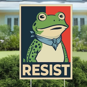 May include: A yard sign featuring a green frog wearing a blue bandana, set against a navy blue and red background. The word "RESIST" is printed in bold, beige letters at the bottom of the sign. The sign is displayed outdoors in a grassy area.