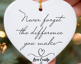 May include: White heart-shaped ornament with gold string, held by a person. The ornament has the text "Never forget the difference you make" in black script, with a heart and "Love Emily" below. The background is a blurred Christmas tree.