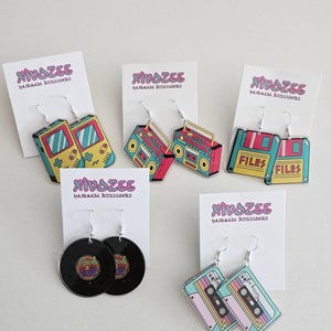 Puede incluir: Una colección de pendientes de temática retro, que incluye una consola de juegos portátil, un radiocasete, un disquete con la palabra "FILES", un disco de vinilo con "Rock & Roll" y una cinta de cassette con "be happy". Cada par se muestra en una tarjeta blanca.