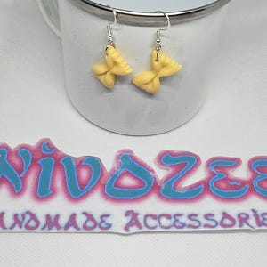 Pode incluir: Um par de brincos amarelos em forma de laço com ganchos prateados. Os brincos são exibidos em frente a uma caneca branca. A imagem também mostra um adesivo com o texto "NIVOZEE HANDMADE ACCESSORIES" em azul e rosa.