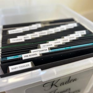 May include: A clear plastic storage bin filled with black file folders labeled for different school grades, from Baby-Toddler to 7th Grade. The bin has a label that reads "Kaden James Keepsakes."