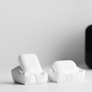 Peut inclure: Deux étuis blancs pour AirPods, chacun ressemblant à une petite chaise rembourrée. Un étui est ouvert, révélant les AirPods, tandis que l'autre est fermé. L'arrière-plan est blanc, avec un haut-parleur noir texturé et une plante visible.