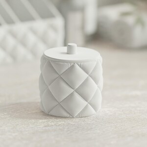 May include: A white, cylindrical container with a quilted diamond pattern and a flat, round lid. The lid has a small, cylindrical handle. The container is likely used for storage or decoration. The overall aesthetic is minimalist and elegant.