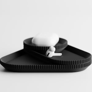 May include: A black, tiered tray with a textured rim, holding a white charging case and a pair of white wireless earbuds. The tray is triangular and sits on a white background. The earbuds are resting on the tray.