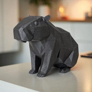 May include: A black geometric capybara sculpture. The sculpture is made of a dark, textured material and features a faceted design. The capybara is sitting upright, with its legs and body clearly defined. The sculpture is on a light-coloured surface.