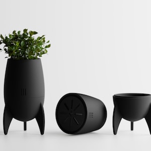 May include: Three black rocket-shaped planters. One upright, filled with green foliage, and two others, one on its side and the other upright. The planters have a matte finish and are designed with three legs and fins.