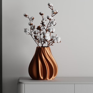 May include: A copper-colored vase with a swirling design holds cotton stems with fluffy white blooms. The vase sits on a white surface against a neutral background, creating a simple yet elegant display.