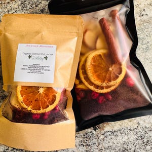 Puede incluir: Dos paquetes sellados de ingredientes orgánicos para ollas a fuego lento. Uno está en una bolsa de papel marrón, el otro en una bolsa de plástico negro. Ambos contienen rodajas de naranja secas, arándanos y otras especias. La bolsa marrón tiene una etiqueta que dice "The Ozark Homestead Organic Simmer Pot packet Holiday".