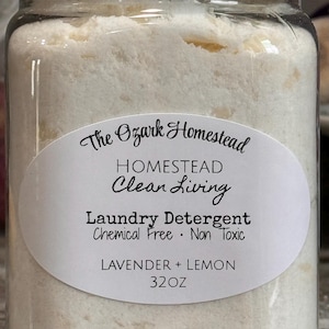 Puede incluir: Un tarro de vidrio transparente lleno de detergente blanco. El tarro tiene una tapa plateada y una etiqueta blanca con el texto "The Ozark Homestead, Laundry Detergent, Lavender + Lemon, 907g". El detergente es libre de químicos y no tóxico.