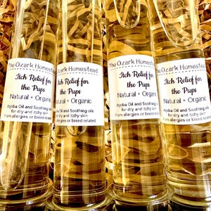 May include: Four glass vials filled with a light amber liquid and brown paper strips. Each vial has a white label with black text that reads "Itch Relief for the Pups" and "Natural + Organic". The vials are on a bed of brown paper.