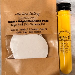 May include: A brown paper package with the text "The Face Factory" and "Clear + Bright Cleansing Pads". Inside the package are white circular pads. Next to the package is a clear tube filled with a yellow liquid.