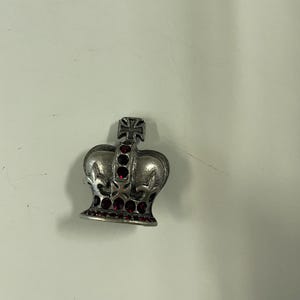 Pode incluir: Um alfinete prateado em forma de coroa com uma cruz no topo e pedras preciosas vermelhas. O alfinete tem um design detalhado e é provavelmente feito de metal. As pedras preciosas são colocadas ao redor da base e no centro da coroa.