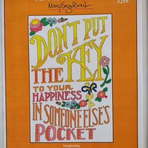 May include: A framed cross-stitch artwork with an orange border. The artwork features the text "DON'T PUT THE KEY TO YOUR HAPPINESS IN SOMEONE ELSE'S POCKET" in yellow, pink, and orange. Floral and key motifs decorate the design.