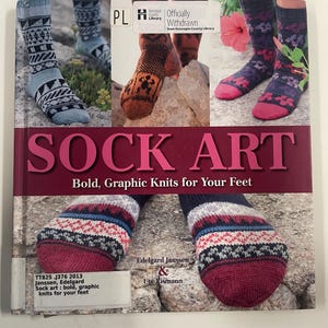 May include: A book titled SOCK ART: Bold, Graphic Knits for Your Feet by Edelgard Janssen & Ute Tismann. The cover features multiple photographs of patterned knit socks in various colors, including blue, brown, purple, and pink, displayed on stone.