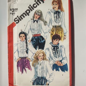 Op de afbeelding: Vintage Simplicity naaipatroon, maat 8 Miss, met illustraties van blouses met ruches en strikken. De omslag is rood en wit, met het nummer 5670 en het Simplicity-logo.