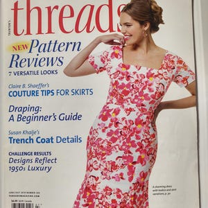 Può includere: La copertina della rivista "Threads" presenta una donna con un abito bianco con un motivo floreale rosa. Il titolo della rivista è in grandi lettere rosse. La copertina include anche testo su recensioni di modelli, consigli di couture e dettagli di trench coat.