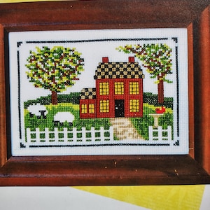 Könnte beinhalten: Ein gerahmtes Kreuzstichbild, das ein rotes Haus mit gelben Fenstern, einer schwarzen Tür und einem schwarzen und braunen Dach darstellt. Die Szene umfasst grüne Bäume, weiße Schafe und einen weißen Zaun. Der Rahmen ist braun. Die Größe beträgt 10 cm x 15 cm.