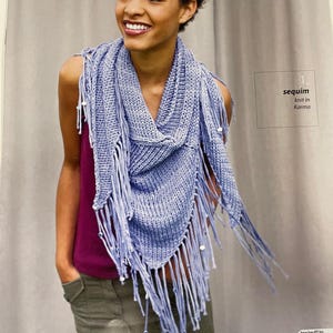 May include: A light blue sequin knit scarf with fringe, part of the Berroco Karma collection. The scarf is draped around the neck and shoulders. The image also includes the text "sequim knit in Karma".