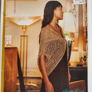 May include: A woman wearing a gold-colored knitted shawl over a dark dress. The image is from a book cover titled "norah gaughan COLLECTION" with the words "objet d'art" at the bottom. The background includes lamps and furniture.