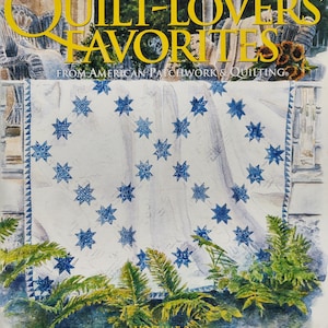 May include: A book cover for "Quilt-Lovers' Favorites" by Better Homes and Gardens. The cover features a watercolor illustration of a quilt with blue star patterns, ferns, and the text "Quilt-Lovers' Favorites" in yellow. The volume number is 2.