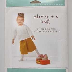 May include: A sewing pattern package featuring a young girl wearing a white long-sleeved shirt and mustard-colored culottes. The package includes the text "oliver + s" and "LUNCH BOX TEE + CULOTTES PATTERN". Sizes are listed as 6-12 M, 12-18 M, 18-24 M, 2T, 3T + 4.