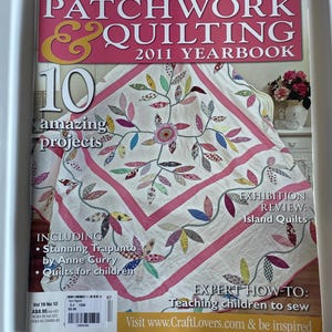May include: The Australian Patchwork & Quilting 2011 Yearbook magazine cover features a white quilt with a colourful floral vine pattern and pink polka-dot borders. Text includes Comprehensive Project Index, 10 amazing projects, and Exhibition Review: Island Quilts, with a barcode and volume information at the bottom.