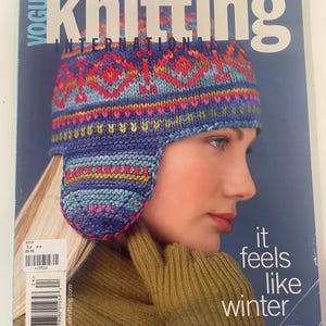 Puede incluir: La edición de invierno de 2002 de la revista Vogue Knitting International presenta una portada con una persona que lleva un gorro de punto con patrones en azul, rojo y amarillo con orejeras y guantes de canalé verde oliva. El texto dice Vogue knitting.