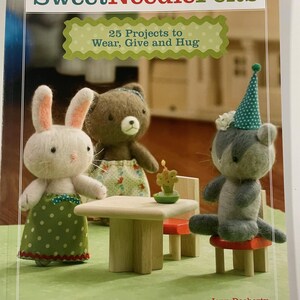Könnte beinhalten: Das Cover des Buches "SweetNeedleFelts" zeigt drei Filztierfiguren: ein weißes Kaninchen, einen braunen Bären und eine graue Katze, die sich um einen Miniaturtisch versammeln. Der Titel ist in Grün und Weiß gehalten, mit dem Untertitel "25 Projekte zum Tragen, Verschenken und Umarmen".