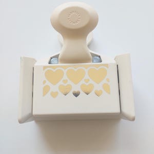 May include: A cream-colored paper punch with a rounded handle and a rectangular base. The base features a gold heart design. The handle is embossed with the text "MARTHA STEWART".