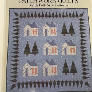 Może przedstawiać: Okładka książki zatytułowana "HOUSES, COTTAGES AND CABINS PATCHWORK QUILTS" z niebieskim wzorem pikowanym. Kołdra przedstawia białe domy z różowymi akcentami i zielonymi drzewami. Nazwisko autora, Nancy J. Martin, znajduje się na dole.