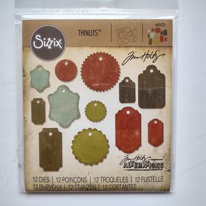 以下が含まれることがあります： 透明なプラスチックパッケージに入った12個のSizzix Thinlits 662423 Tim Holtz Alterations金属製カッティングダイセット。台紙には、赤、緑、茶色の質感のある様々なタグや円形のデザインが印刷されています。