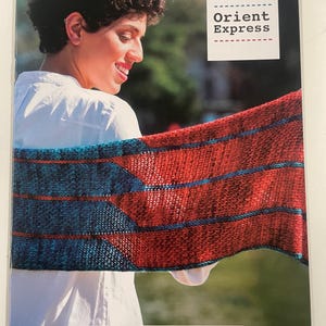 May include: A woman smiles while holding a red and blue knitted shawl. The shawl is made of yarn and has a striped pattern. The image also includes the text "YARN: Mechita Orient Express" and the word "malabrigo".