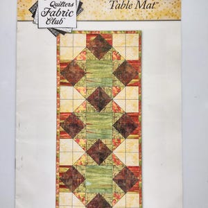 Könnte beinhalten: Rechteckige Tischdecke mit Patchwork-Design. Die Matte hat ein zentrales grünes und gelbes Feld mit braunen Rauten. Auf der Oberseite der Matte steht der Text "Java Gems Table Mat" und das Logo von Creative Quilters Fabric Club.