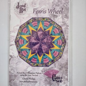 Puede incluir: Un paquete que contiene un patrón "Ferris Wheel" de Jewel Box. El patrón presenta un diseño geométrico colorido con tonos turquesa, amarillo, rosa y morado. El texto "Gems 5&10" y "Cheryl Phillips" también son visibles.