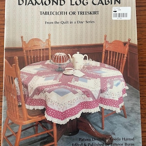 May include: A softcover instructional book titled Diamond Log Cabin Tablecloth or Treeskirt from the Quilt in a Day series. The cover features a round table with a pink, white, and blue patchwork quilt, a cake, and a teapot, designed by Frieda Harrod.