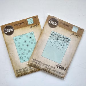 May include: Two Sizzix Texture Fades A2 embossing folders in clear plastic packaging. The left folder features a light blue starburst pattern, while the right folder displays a black speckled gradient design on a light blue background.