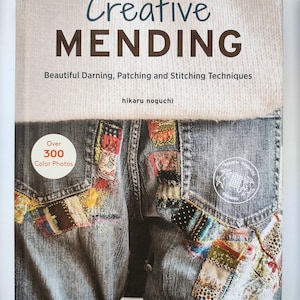 Puede incluir: Portada del libro "Creative Mending" con el subtítulo "Beautiful Darning, Patching and Stitching Techniques". La portada presenta un fondo de mezclilla con parches de tela coloridos. El libro tiene más de 300 fotos a color.
