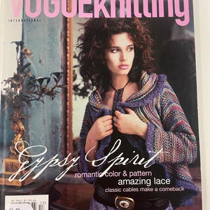 May include: Vogue Knitting International magazine cover featuring a woman wearing a knitted jacket. The cover has the text "Vogue Knitting" in pink, with "Gypsy Spirit" and other text. The jacket is purple, blue, and multicolored.