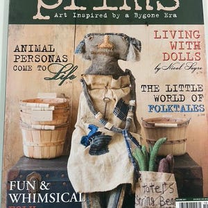 Może przedstawiać: Okładka magazynu z lalką ludową w beżowej lnianej sukience i dżinsowych akcentach. Tytuł magazynu to "prims" z podtytułem "Art Inspired by a Bygone Era". Inne teksty to "Animal Personas Come to Life" i "Fun & Whimsical Folk Figures."