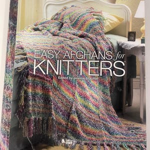May include: A softcover book titled Easy Afghans for Knitters edited by Jeanne Stauffer, published by House of White Birches. The cover features a photograph of a multicolored, fringed knit afghan draped over a bed in a bedroom setting.