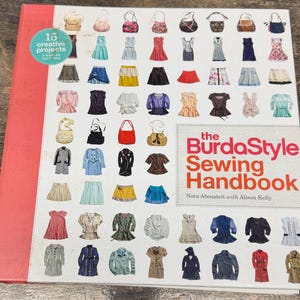 Pode incluir: O livro The BurdaStyle Sewing Handbook de Nora Abousteit com Alison Kelly tem uma lombada vermelha e uma capa branca com uma grade de várias peças de vestuário e bolsas ilustradas, além de um círculo azul-petróleo com 15 creative projects.