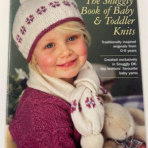 May include: A book cover featuring a young child wearing a knitted hat, scarf, and sweater. The hat and scarf are cream-colored with a burgundy trim and floral accents. The sweater is a deep magenta color. The book title reads "The Snuggly Book of Baby & Toddler Knits."